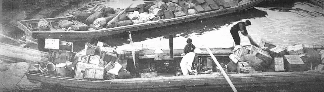 Sepia-toned photograph taken in about 1910 of three men in a loaded scow on a river bank arranging boxes to balance the load.