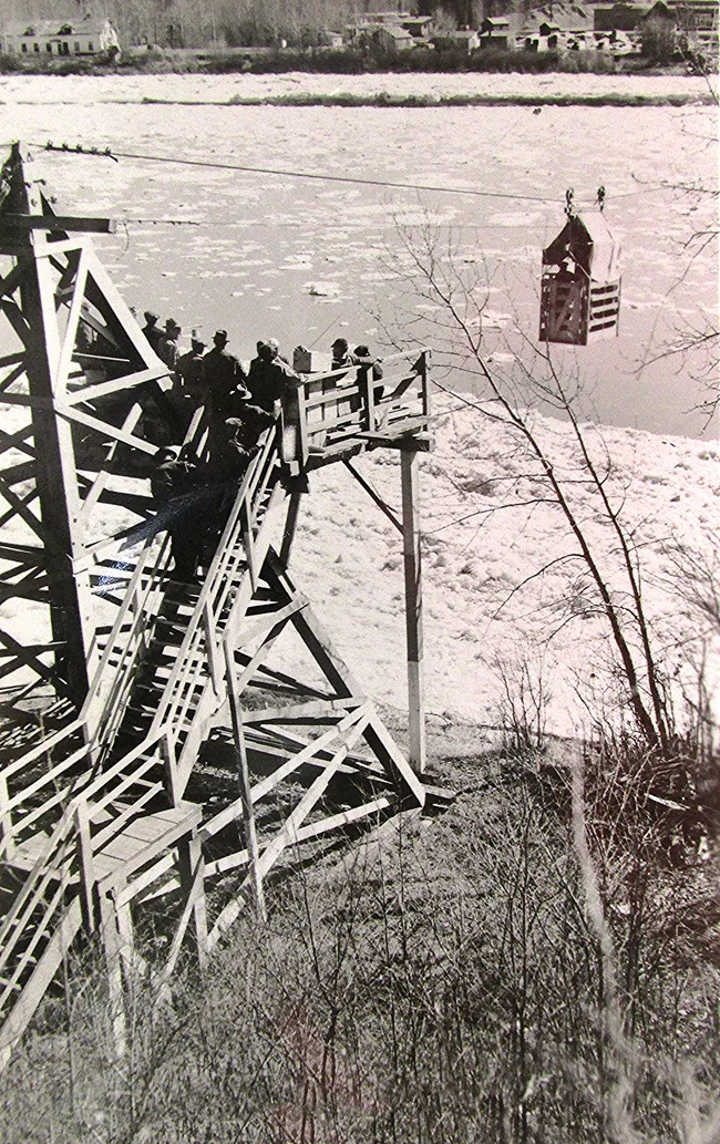 Black and white photo of a wood-framed tower on the bank of a river. It is spring time and the ice is defrosting from the river. suspended from the tower is a cable with a wood-framed cage hanging from it on two wheels with a pully system. There are people queued for passage, standing stairs and a platform built onto the tower.