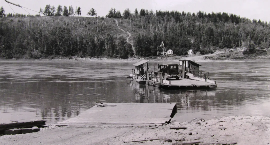 A wooden ferry floating away from a dock on a river carrying a Ford Model T and with shacks built on the sides of the deck.