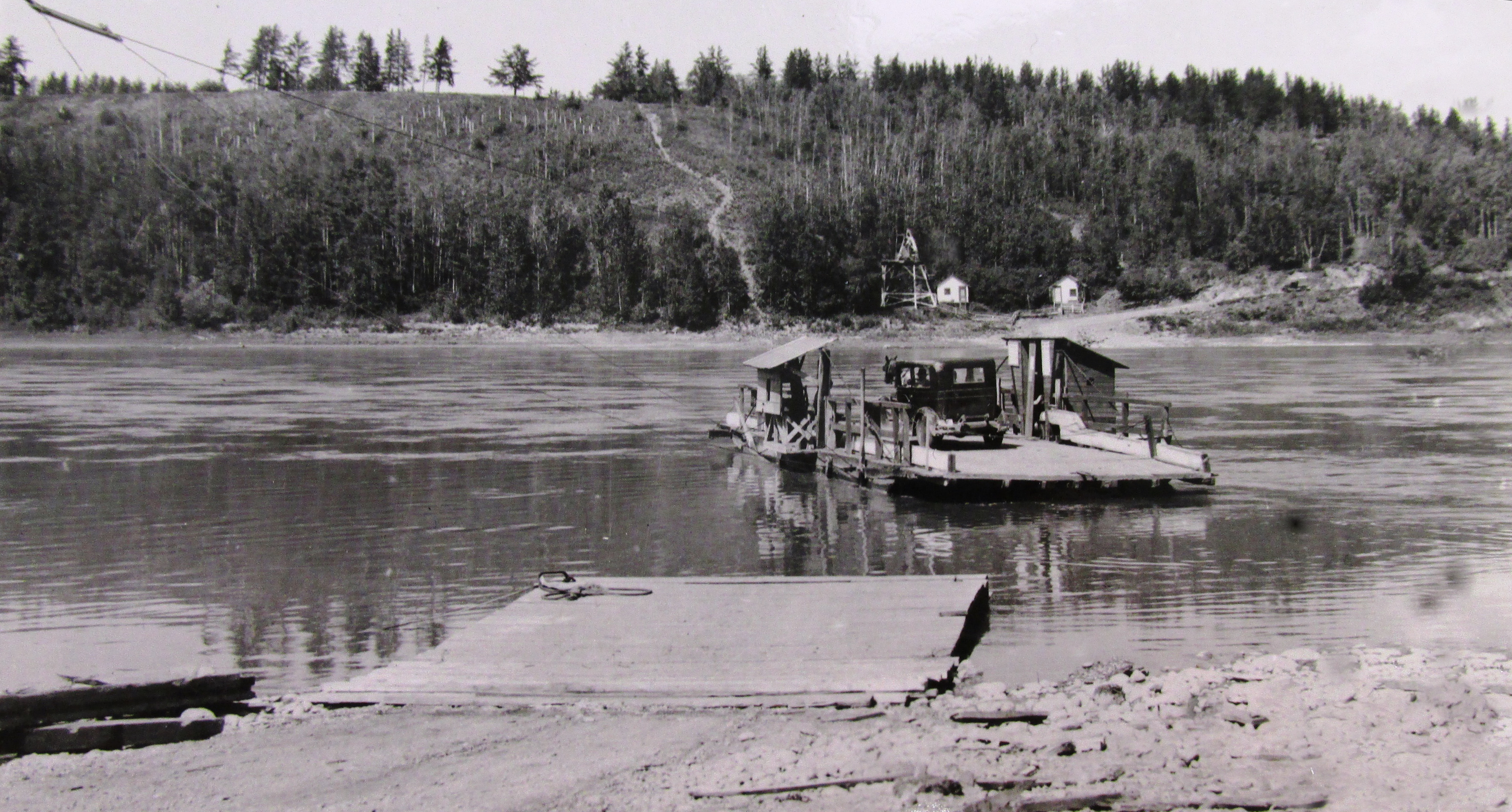 Black and white photo taken circa 1920s of a car on the deckof a wooden ferry that is floating across a river. The opposite bank is heavily wooded and a trail and a track can be seen leading down to the landing. On the near shore is a wooden landing in the water off the river bank.