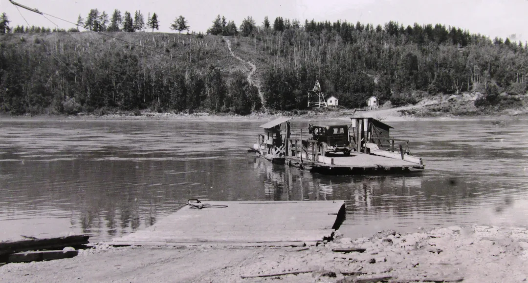 Black and white photo taken circa 1920s of a car on the deckof a wooden ferry that is floating across a river. The opposite bank is heavily wooded and a trail and a track can be seen leading down to the landing. On the near shore is a wooden landing in the water off the river bank.