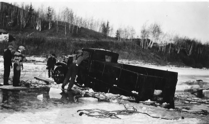 Black and photograph of a 1930s-era panel truck half submerged in an icy river. A man is standing on a block of ice, reaching into the cab. Four other men are standing on the ice, looking on and holding cables and ropes.