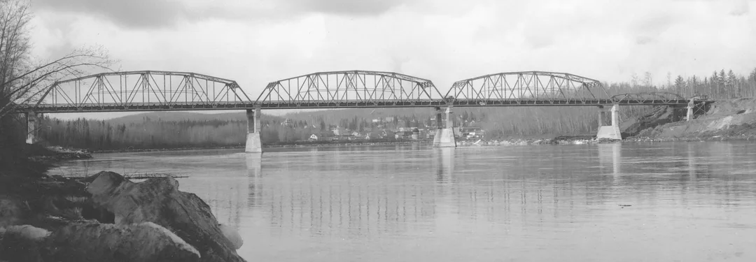 Black and white photograph taken in 1952 of a truss-construction bridge crossing over a river. It is flanked by rocks on the shore and trees, and a small town can be seen across the river, framed by the bridge, in the distance.
