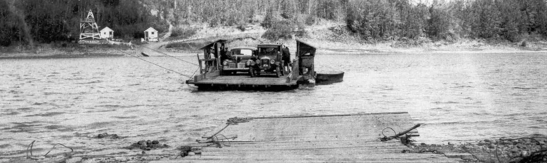 Black and white photo of two old cars vintage 1920s to 1930s on the deck of a wooden ferry which is attached to cables and is floating across a river.