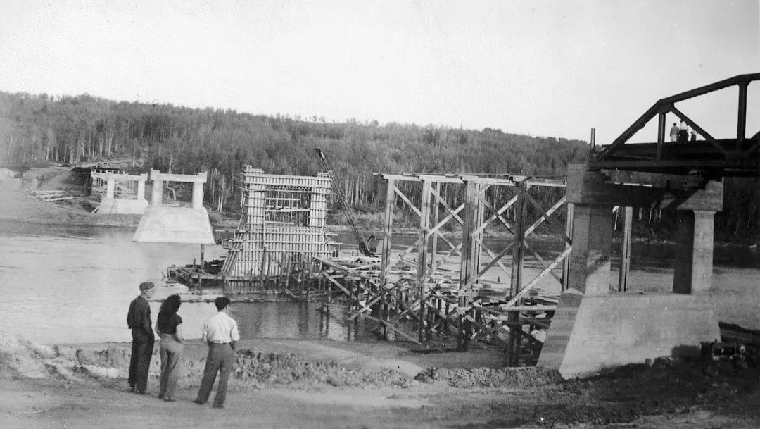 Black and white photograph taken in the early 1950s of three people standing on a river bank looking at a bridge under construction on a wide river. The are pilons in three stages on constuction; framing, metal rebar framing and poured concrete. There is no deck on the bridge.