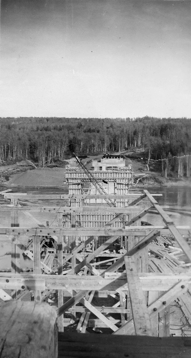 Black and white photograph of a bridge under construction across a river.