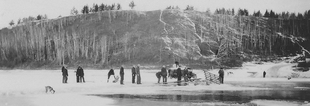 Groups of people standing on a frozen river with the wooden panels of a truck sticking up out of the water.