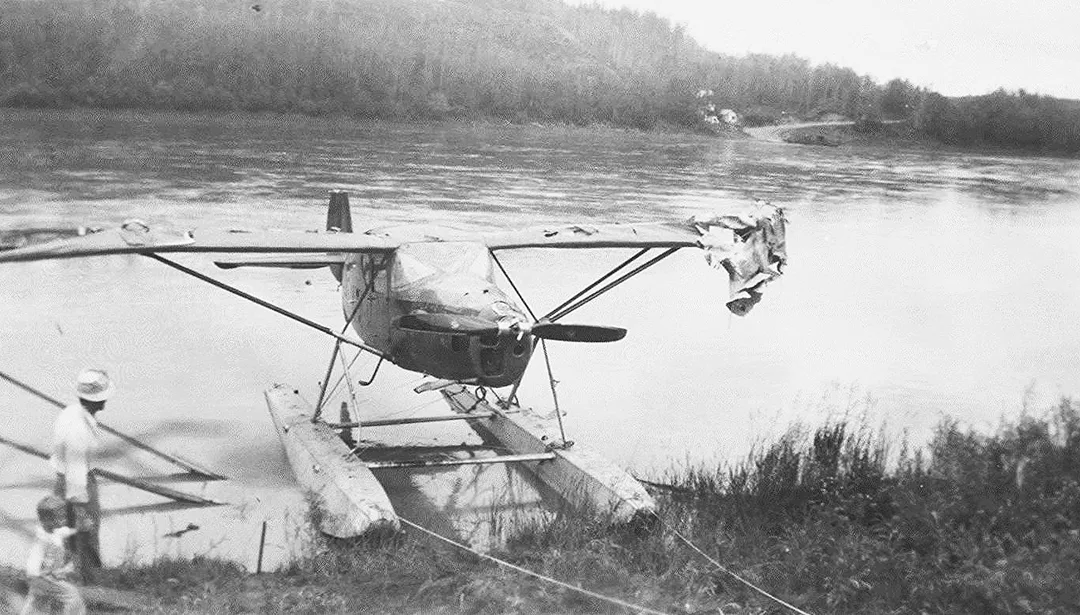 A single-seater, propellor airplane on floats landed on a river bank showing damamge to one wing. A man holding the hand of a little boy are approaching the plane on the riverbank.