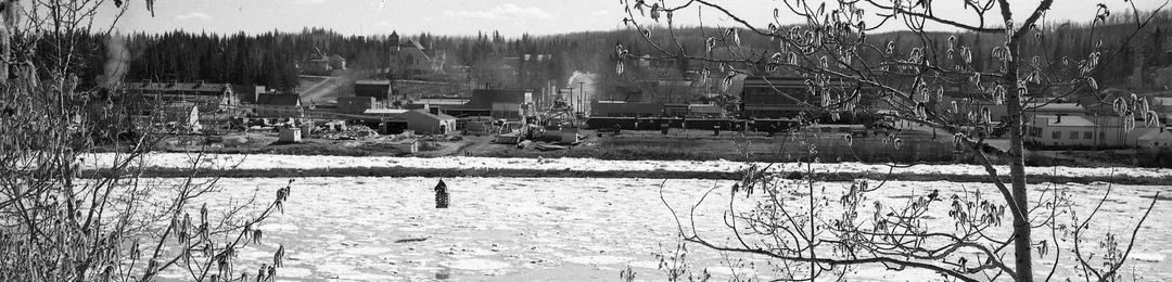 Black and white photograph of a partially defrosted river with broken ice blocks flowing in it. A small town can be see on the opposite bank. There is a small, wood-framed cage over the water, suspended from a cable spanning the river.