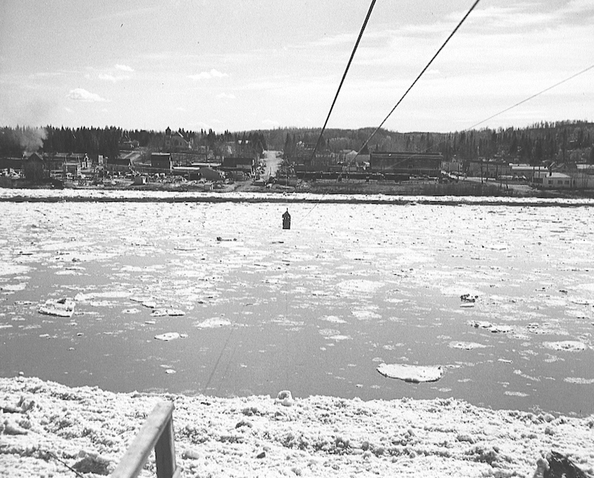 Black and white photo of a small wood-framed cage suspended from a cable over a river with broken up ice floating in it. There is a small town built up on the opposite bank of the river.