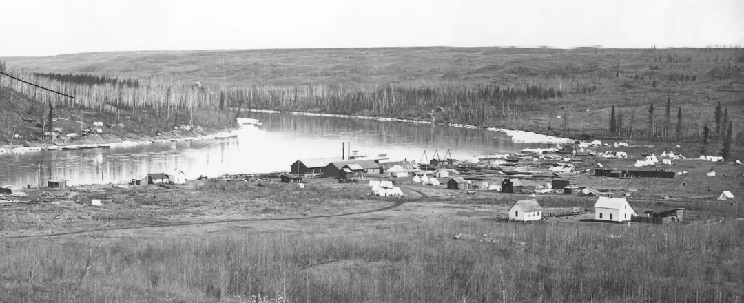 Photograph of buildings and boats under construction on a broad, flat bank at the bend of a river. It appears to be spring with ice on the banks of the river and no leaves on the trees.